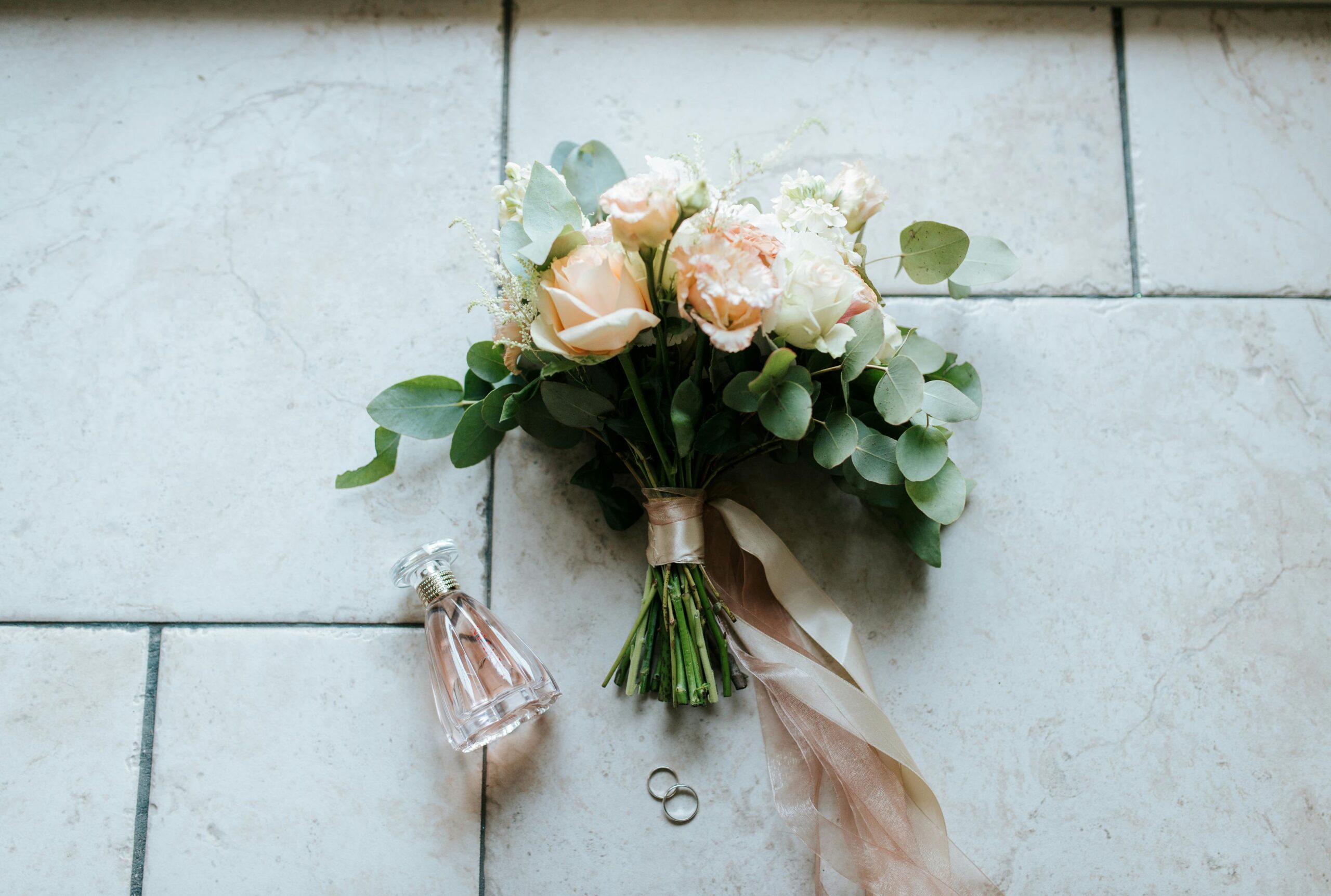 Beautiful wedding bouquet with roses, rings, and perfume bottle arranged on a tiled floor.